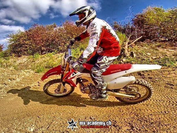 Dirt bike riding standing up: a rider on a red Honda in the corner in front of reddish-brown bushes