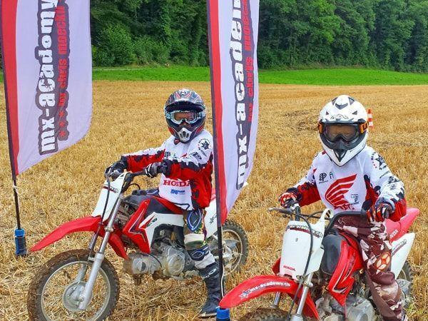 Kids dirt bike: two children sitting on Honda kids' motorbikes, with MX-Academy flags flying left and right