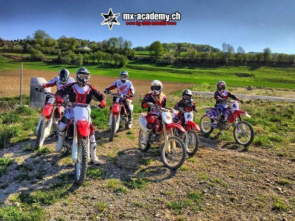 Motocross events: a group of six riders with their bikes on gravel, green hills in the background