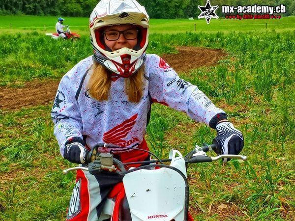 Motocross pictures: female rider laughing under a white helmet with goggles, sitting on a Honda, greenery all around