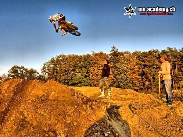 Motorbike riding with an audience: a rider jumps high over a dirt mound, two spectators stand on top